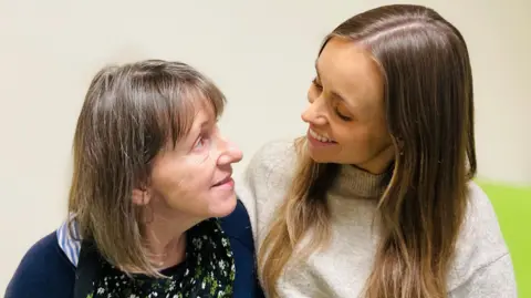 BBC Patient Samantha Denison is sitting on a hospital bed and looking at her daughter Steph. Samantha has shoulder length brown hair and wears a dark blue cardigan over a floral top. Steph has long brown hair and wears a light grey, high-neck jumper.