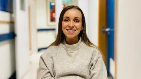 Steph Denison is sitting in the hospital ward. Wearing a soft grey jumper, she is smiling.