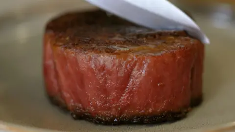 BBC A sirloin steak, well cooked. Dark on the top and pink around the sides. A knife begins to cut it