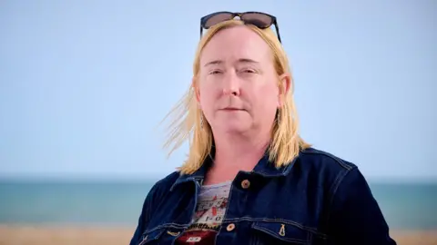Jim Holden / BBC A woman with blond shoulder-length hair, standing on a pebble beach with the sea in the background. She is wearing a blue denim jacket. The sky is blue and cloudless.