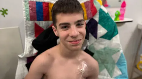Steve Hubbard/BBC Finley sitting upright in a hospital bed. He has a medical tube attached to his chest. He is smiling at the camera. On the bed behind him is a multi-coloured patchwork blanket. Finley has dark brown straight hair.