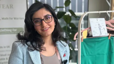 BBC Roya Rasouli is smiling directly at the camera. She is standing next to a collection of leggings designed to help those who struggle with endometriosis. She is wearing a light blue blazer and has a tape measure around her neck. 