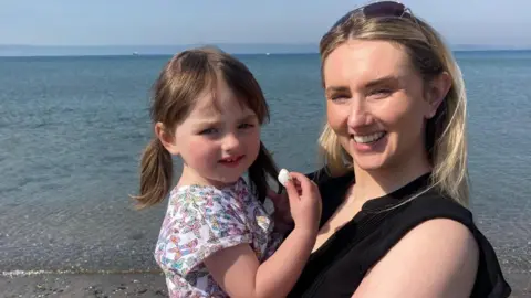 BBC Shelley and Ruby smile as they stand on the shoreline at the beach looking at the camera. Ruby is in Shelley's arms and is eating a marshmallow. She has brown hair in a double ponytail and is wearing a white t-shirt with colourful patterns on it. Shelley has naturally dark hair which has been dyed blonde. She has sunglasses propped on top of her head and is wearing a black shirt. Shallow water is visible behind them, which stretches out to the mountains on the horizon.
