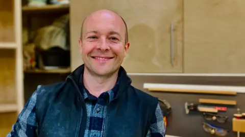 BBC Furniture maker Nick James is in his furniture making workshop in Newcastle. He is looking at the camera and smiling and wearing his casual clothing. Behind him you can see his wood-working tools on the wall.