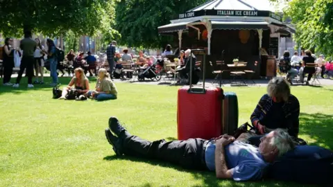 PA Media A man reclines in the shade on grass in front of a pavilion selling ice cream in Edinburgh's Princes Street Gardens.