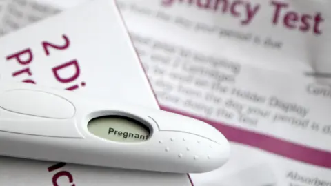 PA Media A positive pregnancy test lies on top of the test box which is white with magenta writing, the instructions for the kit are blurred out in the background.