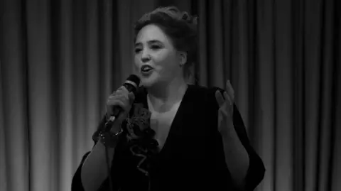 Scary Little Girls A black and white photo. A woman wearing a black dress. She is holding a microphone in her hand. She has her hair up in a bun. There are curtains behind her. 