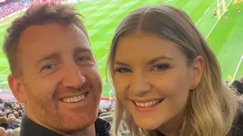 Siobhan Gorman A selfie of Siobhan and her partner who are in a rugby stadium. Both are smiling at the camera from the seats with the pitch behind them. She has long wavy blonde hair and is smiling. He has short ginger hair and a cropped beard.