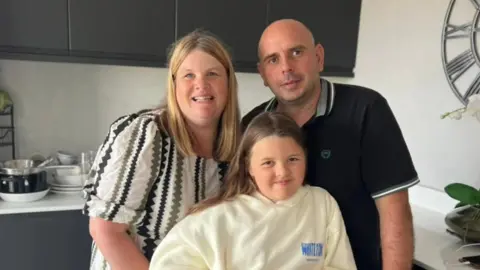 Jenni Whitmore Jenni with her husband Gareth and young daughter Carys in their kitchen. Jenni has her blonde hair down and is wearing a white, black and green dress. Her husband is wearing a black polo shirt. Her daughter is wearing a yellow hoodie and has long brown hair.
