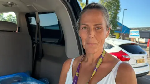 BBC A woman in a white vest standing at the open boot of a van in a car park looking at the camera