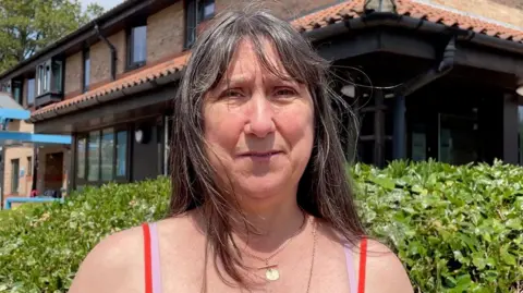 Kit Taylor/BBC A woman with brown hair looking directly into the camera, with a serious expression. She is wearing a bright orange vest with straps and two golden necklaces.