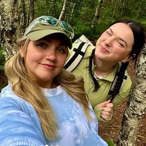 Emily & Lucy Emily and Lucy - two women standing in a forested area, surrounded by trees. One is wearing a light blue tie-dye shirt and has long blonde hair. The other is dressed in a green shirt and carrying a backpack.