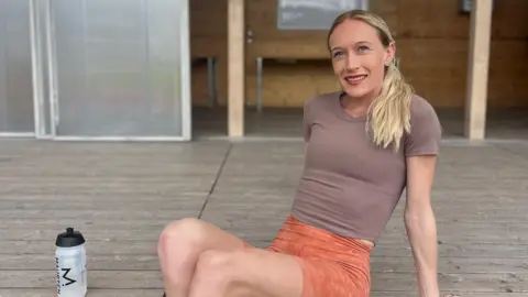 Calli Hauger-Thackery Calli with the slender frame of a distance runner, blonde hair over one shoulder, blue eyes, red lipstick and sat on wooden decking leaning back on her hands.