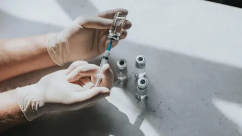 Getty Images A gloved medical professionals' hand uses a syringe to extract liquid from some vials on a table. 
