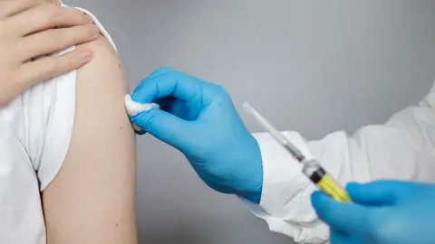 Getty Images A person lifts up their sleeve as a medical professional wearing gloves holds a pieces of cotton wool to their arm while they hold a syringe in their other hand