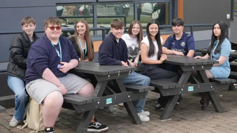 BBC A group of male and female students, all aged 17, sit together, smiling, at two picnic tables in front of their college building