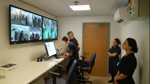 Hospital staff are using a microphone to give information to people in the lab about the unfolding emergency scenario. Two women are standing watching the screens showing what is happening in the lab.