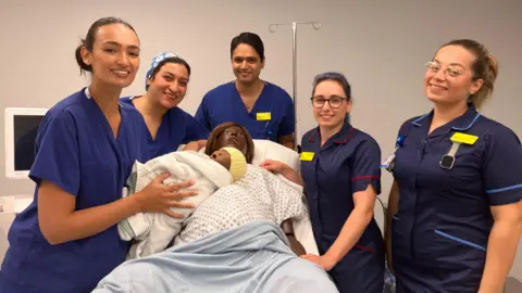 BBC Staff using the simulation lab are standing around Linda the mannequin who has just given birth. The maternity staff are wearing blue uniforms with yellow name badges and holding a doll baby wrapped in a blanket and with a knitted yellow hat on. There is equipment around them, including a screen.