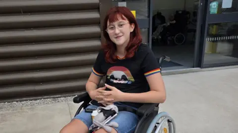 Family handout Wren is smiling into the camera. We see her in her manual wheelchair. She is wearing a blacktop with rainbow edging on the short sleeves and blue denim shorts. Wren has dark red hair and is wearing glasses. 