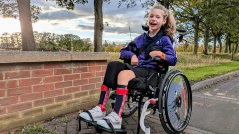 Family handout Wren is looking directly into the camera with a wide smile. She is wearing her school uniform and is sitting in her manual wheelchair. Wren's hair is tied up in a ponytail and is wearing leg splints and white trainers.
The picture has been taken in a wooded area. 