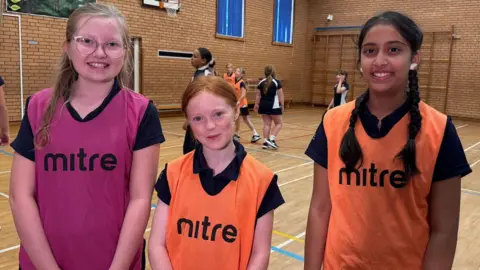 Three Year Seven girls are in a school sport hall. They are smiling at the camera. They are wearing navy short-sleeved uniform T-shirts and coloured netball bibs. A game of netball is taking place in the background.