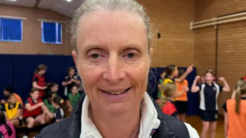 Liz Brown is wearing a white sports top and navy Gillet. She's smiling at the camera. She's in a school sports hall and there are students laughing behind her.