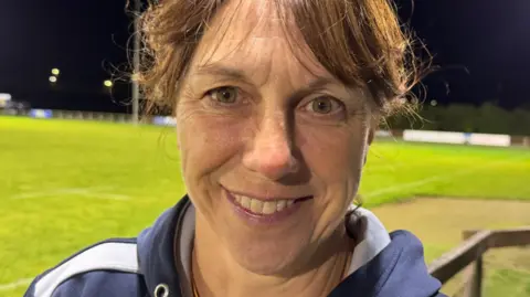 Nicky Crawford is standing next to a floodlit rugby pitch. She's wearing a hooded training top and smiling.