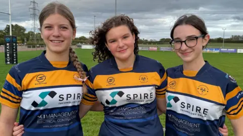 BBC The three teenagers are standing on a rugby pitch with their arms around each other. They are smiling and wearing blue and gold Worcester rugby club shirts.