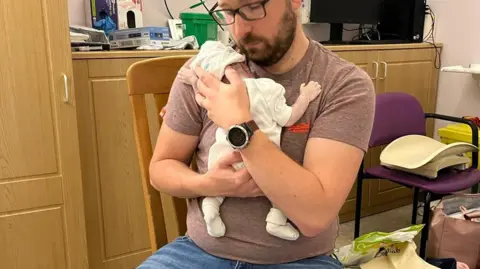 Family picture A man with beard and glasses, wearing jeans and a t-shirt, is sitting on a chair in a hospital side room. He holds to his chest an infant, who is dressed in white baby clothes. Cupping the baby's head, the man's face is brushing the baby's. The man has a stoic expression.