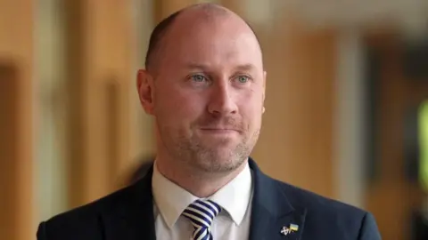 PA Media Neil Gray, a bald man with ginger stubble, looks off camera to his left. He is wearing a navy suit jacket, white shirt and striped tie