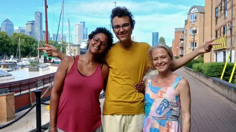 Tunde, Buddy and Kellie stand in a row on a path by a river. Boats can be seen along the river beside them and there are skyscrapers in the distance.
Tunde has short, dark curly hair and is wearing a red strappy top with grey trousers. She has her arm around Buddy's waist. Buddy has dark curly hair and glasses. He is wearing a yellow t-shirt with beige shorts and has his arms outstretched with his index fingers pointing. Kellie has blonde hair and is wearing a colourful patterned summer dress with an abstract design.