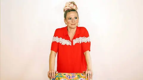 Kellie Bright Kellie Bright is wearing a bright red shirt with white lace details, leaning forward with hands on a colourful floral-patterned surface, against a plain light background. Her hair is tied up with a pink accessory.