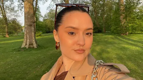 BBC A selfie of a young woman in a park. She has brown hair pulled back and tortoiseshell sunglasses on her head. She has a light brown mac on and dark brown hooped earrings. Trees and grass are in the background.