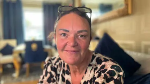 Kellie Howson is sitting in her living room. She has dark hair scraped back into a ponytail, and sunglasses on the top of her head. She is wearing a leopard print-style blouse. 