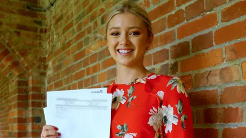 PA Media A young woman with blonde hair tied back. She is smiling at the camera and is stood in front of a brown brick wall