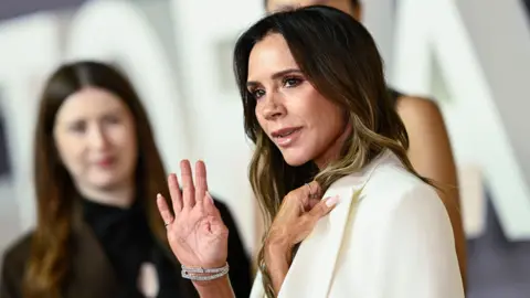 Getty Images Victoria Beckham waves while wearing a white suit with other people in the background as she attends the Victoria Beckham premiere in London on Wednesday. 
