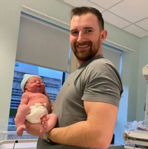 Julie Keegan Angus Mason who is in a grey T-shirt, holds a newborn baby in a hospital room