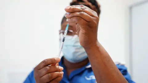 PA Media A close up shot shows a nurse holding a syringe inside a vile while preparing a Covid booster jab, she's wearing a medical facemask and blue nurse outfit against a white background. Picture taken at University Hospital Coventry in 2022.