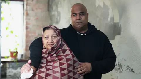 Nick Woolley/BBC Bushra and Atif standing close together indoors against a wall with peeling paint and visible damage. Atif is wearing a dark hooded sweatshirt and has an arm around Bushra, who is wrapped in a patterned shawl with red and beige triangles. A window with a potted plant is partially visible in the background.