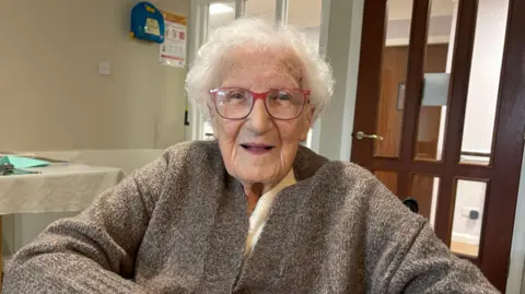 BBC An elderly woman, 100-year-old May O'Shea, smiles into the camera. She has short, white curly hair, blue eyes, red glasses and is wearing a brown cardigan.