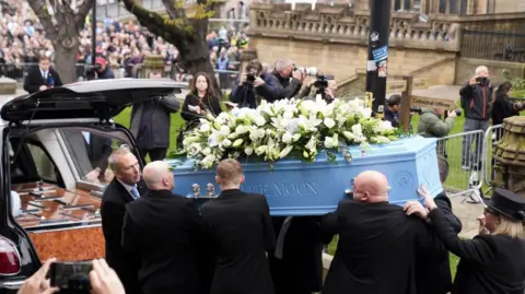 PA Media A blue coffin, which is adorned with white flowers, is carried out of Manchester Cathedral by Ricky Hatton's family and put into the back of a hearse. Crowds of people can be seen paying their respects in the background. 