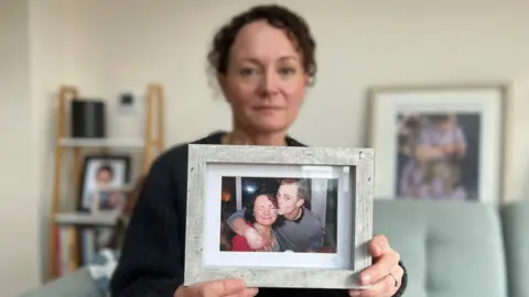 Stuart Woodward/BBC Emma Harley holds up a framed photograph of her and her brother Terry White. Emma has dark curly hair, she is wearing a dark cardigan, and she is sitting on a green sofa surrounded by other photographs. In the photograph she is holding, she is wearing a red top and grimacing at the camera with her eyes closed. Her brother Terry has an arm around her shoulder and is giving her a kiss on the cheek. He has short shaved hair and is wearing a grey top.