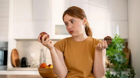 Getty Images Young woman holding an apple and a chocolate cookie, contemplating a healthy food choice in a stylish, modern kitchen