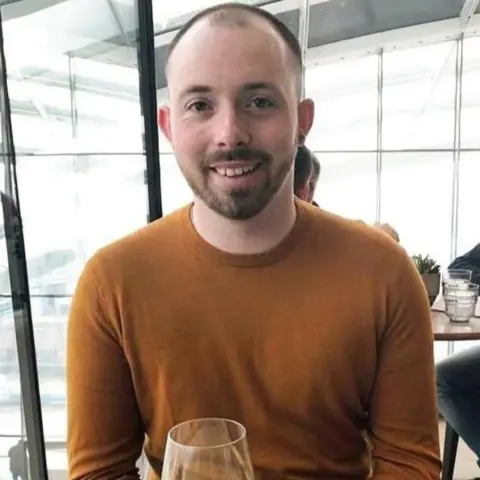 Ian Casey in a brown jumper. His hair is receding and a small island of hair is left on his head.