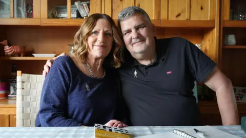 Danny Burkey Man and woman both dressed in blue sitting at a dining table, looking at the camera. There is a book of crosswords and pen and paper on the table. Behind them is a wooden cabinet. 