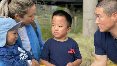 Oliver, smiling, sits on his mother Jingru's knee. He is wearing a blue cap, a blue top and bright yellow trousers. Jingru has long blonde hair in a ponytail and is wearing a white T-shirt and blue cardigan. She has her other arm around her eldest son Skyler who is wearing a blue T-shirt with red writing on it. Dad Ricky is sitting next to Skyler and is wearing a blue T-shirt and yellow trousers. He has very short hair.