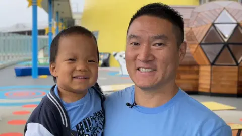 Dad Ricky is carrying Oliver in his arms. They are borth smiling and wearing blue T-shirts. They are on a rooftop with colourful seats and tables in the background.
