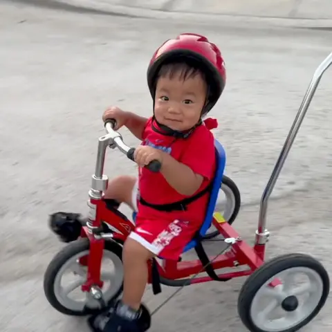Chu family Oliver Chu rides a red tricycle. He is wearing a red helmet, red T-shirt and red and white shorts and is looking at the camera