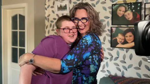 Adam hugs his mother, the Reverend Charlotte Cheshire. Both are smiling into the camera. They are in a living room with family photos on the wall and grey, blue and cream leaf-patterned wallpaper. Adam is on the left and wears a purple t-shirt and red-and-black glasses. His mum is grinning and wears a black dress pattered with purple, green and blue and black and animal-print glasses. She has curly brown hair in a bob.