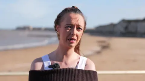 BBC / Jadzia Samuel A blonde woman wearing a white top and black jumper is standing by a beach.